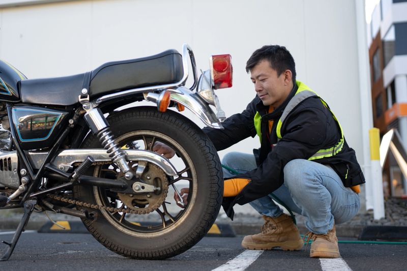 A man performs a visual inspection of his motorcycle's rear tyre in a parking area.