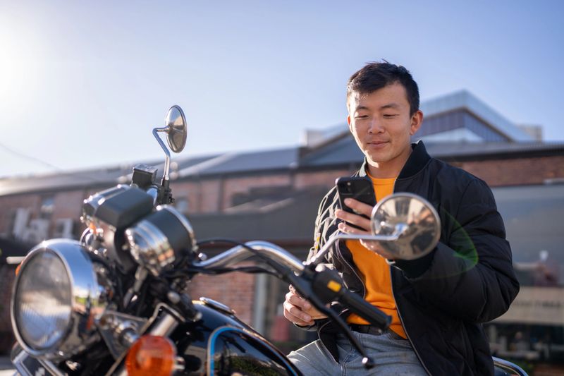 A man sits on a parked motorcycle while looking at his smartphone in an urban setting