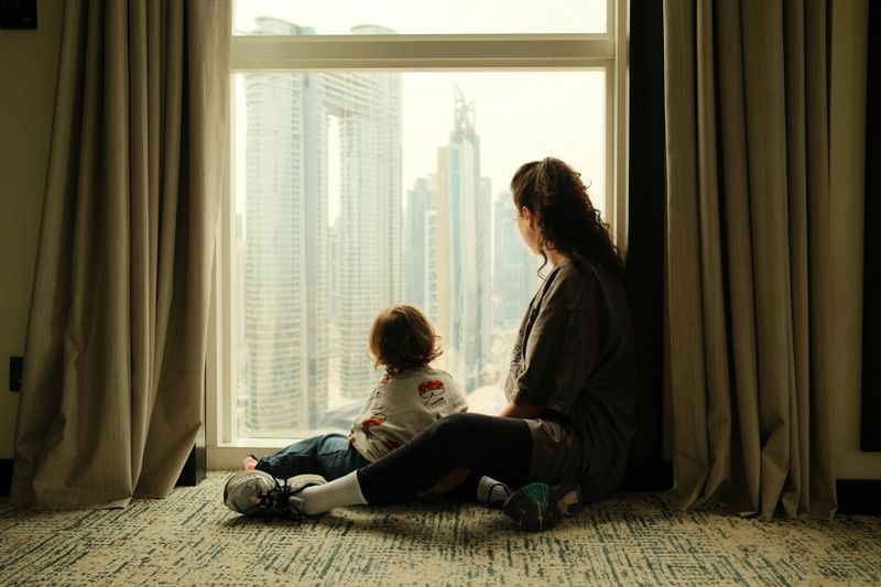 Mother and son sitting on the floor, looking at the Dubai cityscape through the window of their hotel room