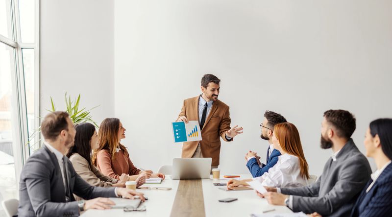 Smiling leader standing at boardroom and showing chart with finances to his leadership team at conference table.