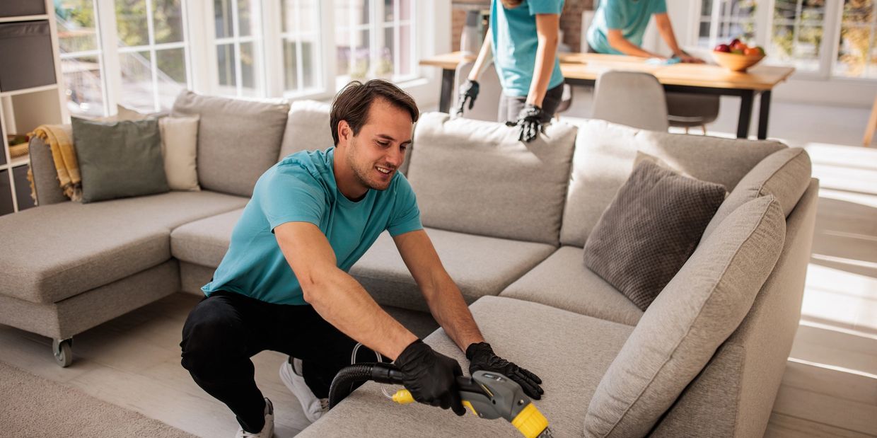 Man cleaning a gray sofa with a handheld steamer in a bright living room.