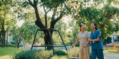 A nurse assists an elderly woman using a walker outdoors in a sunny garden.