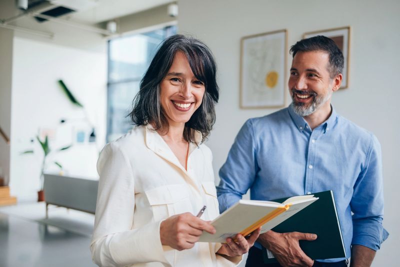 A confident woman holding a notebook and pen, accompanied by a smiling professional male colleague, working together in a bright and modern office environment.