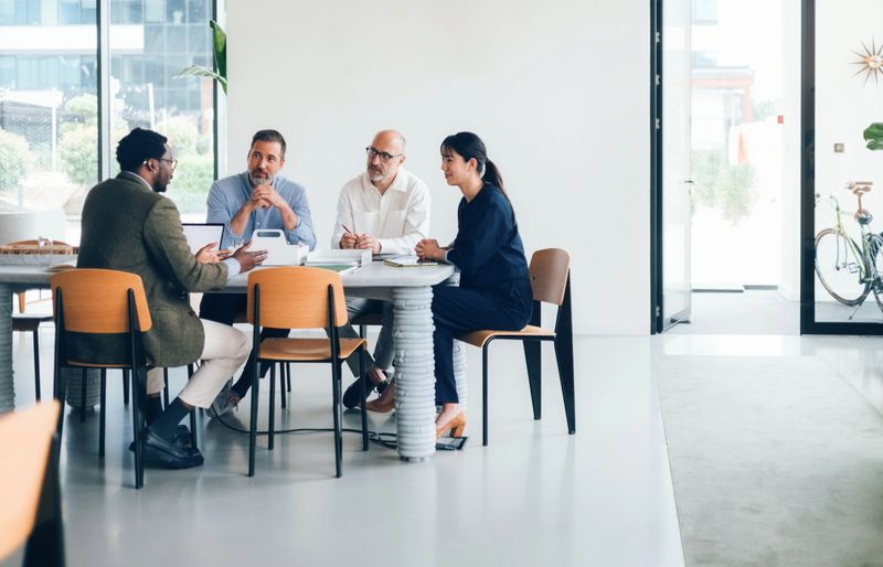 Group of professionals seated at a table discussing ideas, showcasing teamwork and focus in a bright, modern office setting with minimalistic interiors.