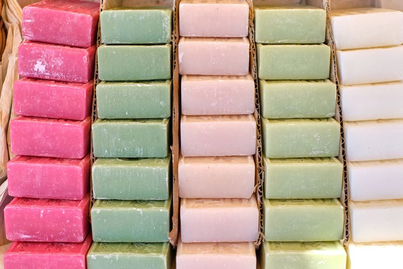 A neat, vibrant arrangement of handcrafted soap bars in soft pastel hues—pink, green, beige, and white—sits on display at a local market, reflecting a commitment to natural skincare and artisanal craftsmanship. The textures and tones evoke cleanliness, self-care, and eco-conscious living.