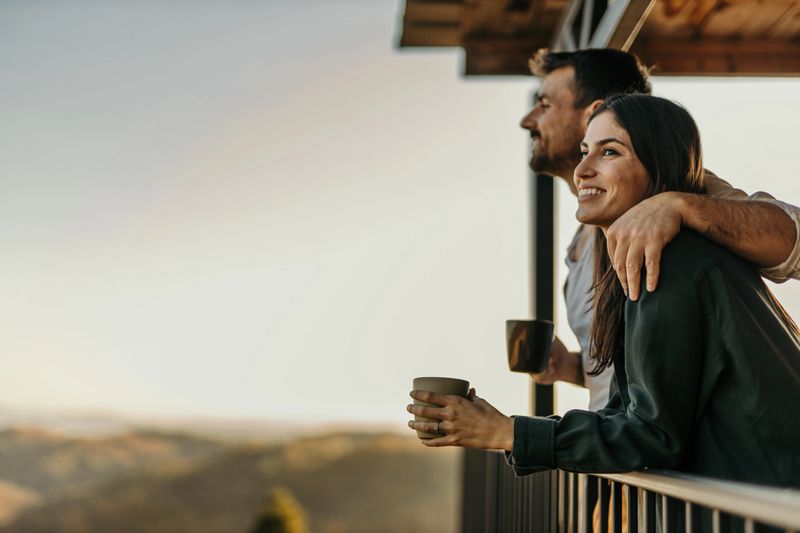 Couple standing on balcony, drinking coffee and enjoying sunrise over scenic landscape