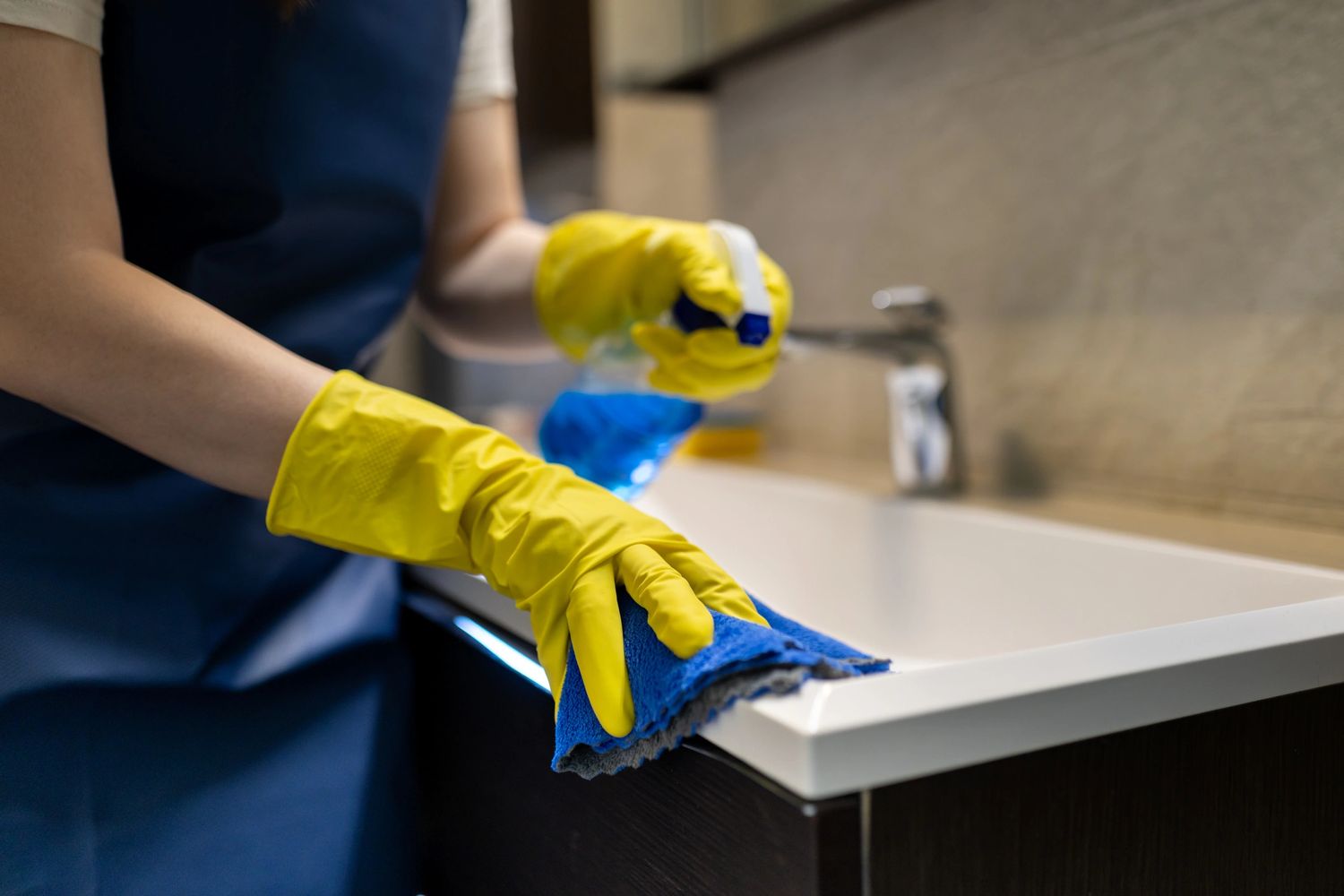 Person cleaning a bathroom sink wearing yellow gloves and using a blue cloth.