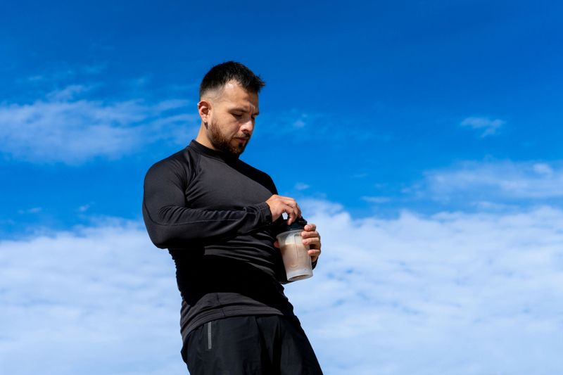 A muscular young man wearing tight black sportswear stands under a vibrant blue sky while preparing a protein shake in a translucent shaker bottle. His focused expression and athletic posture suggest a disciplined workout routine and commitment to sports nutrition
