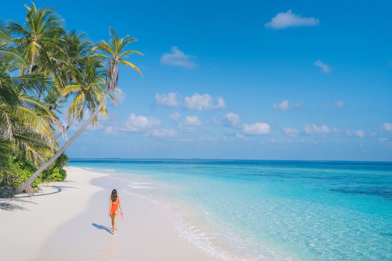 Aerial view of young woman walking on white sand beach along turquoise sea, palm trees