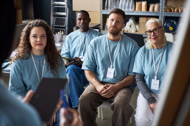 Group of diverse individuals attentively listening to a speaker at workshop. Participants wearing blue shirts and name tags, seated in organized setting with shelves in background