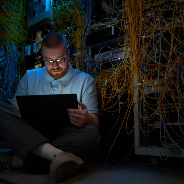 Man working on a laptop in a server room with tangled cables.