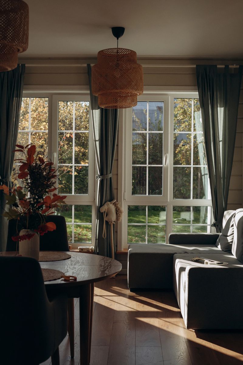 Soft sunlight filters through large windows, illuminating a cozy living room adorned with potted plants and elegant decor. The atmosphere is warm and inviting.