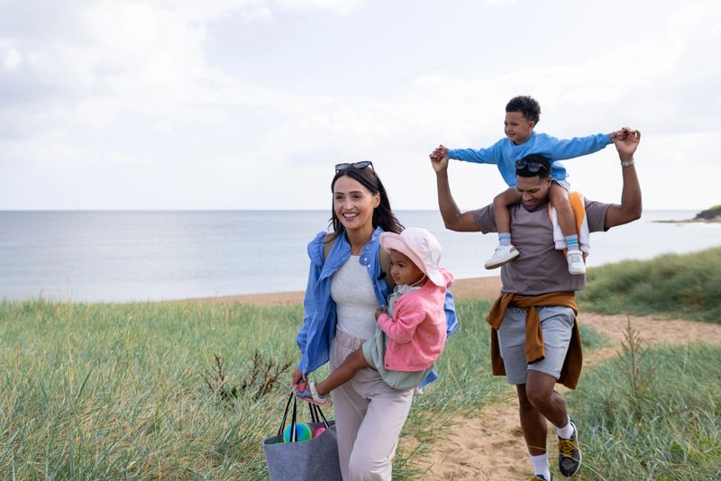 A wide shot of a cheerful young family walking along a sandy path surrounded by grassy dunes. The mother is carrying her toddler daughter while smiling. The father follows closely behind, carrying his son on his shoulder while holding onto his hands. The peaceful coastal backdrop features a stretch of sandy beach and calm ocean waters.

Videos are available similar to this scenario.