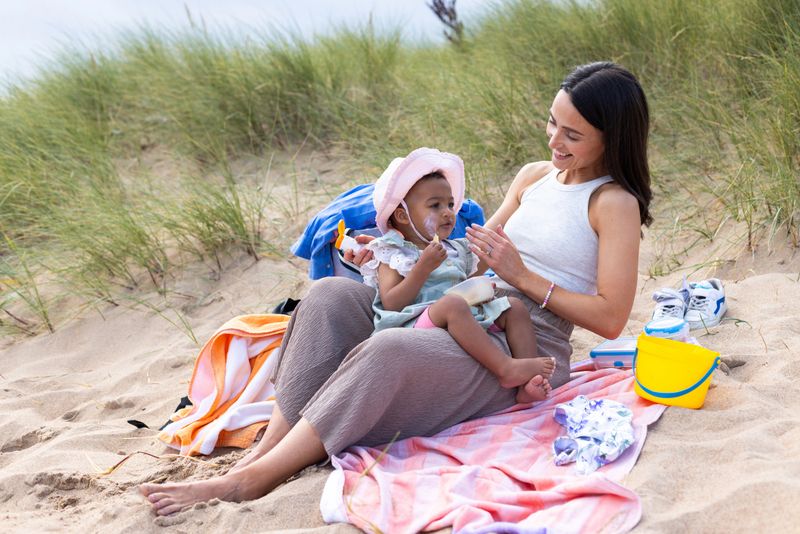 A medium shot of a mother and daughter sitting together on a beach in Seaton Sluice. The daughter is lying on her mother's legs while the mother applies sun cream to her face. They are lying on a towel with sand dunes behind them.Videos are available similar to this scenario.