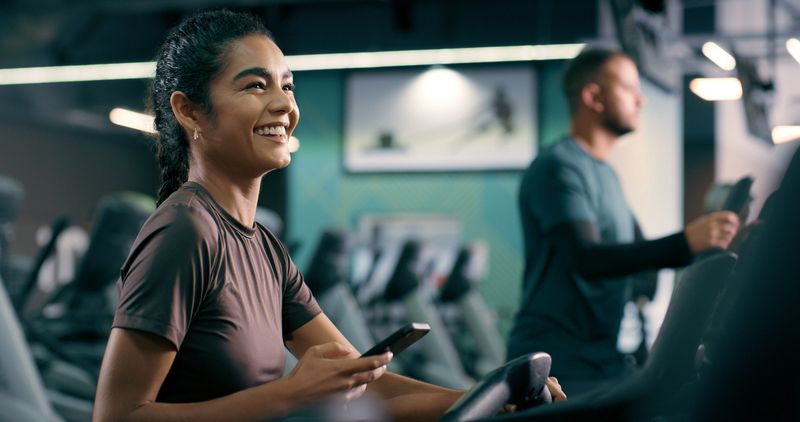 Gym, woman and fitness with smartphone for joke, online communication and cycling with laugh. Machine, female person and chat with cellphone app for exercise meme, virtual workout and social media