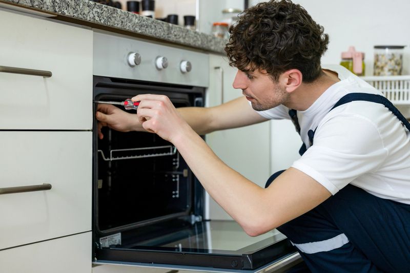 Young repairman in protective workwear fixing oven in kitchen