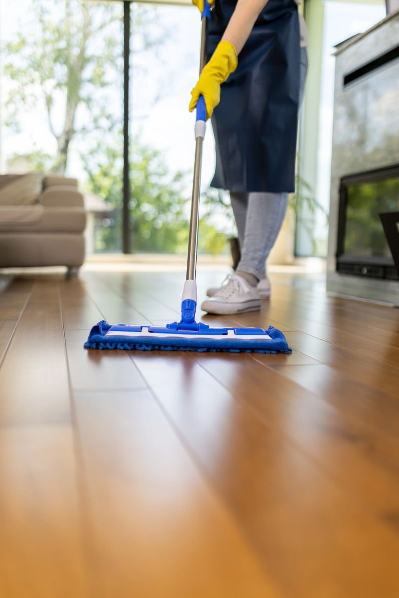 A person in yellow gloves is mopping a hardwood floor in a spacious room with large windows. Natural light streams in, highlighting the clean surfaces and modern decor.