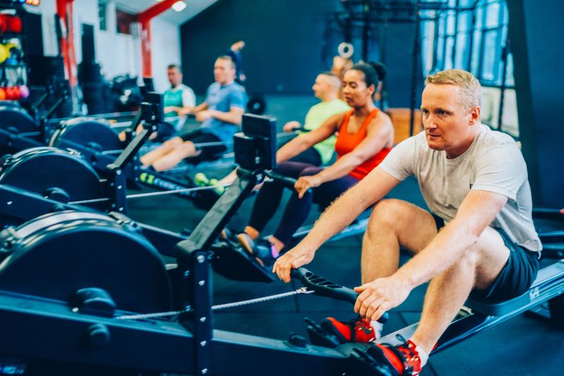 A group of individuals work out together on indoor rowing machines in a modern gym. The energetic atmosphere, bright lighting, and variety of fitness equipment highlight teamwork, motivation, and a commitment to health and wellness.