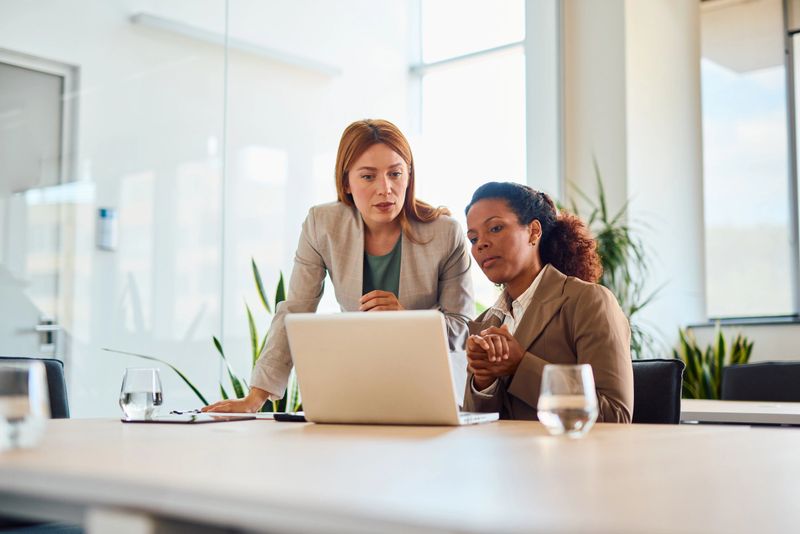 Two focused businesswomen collaborating on a project, analyzing data on a laptop in a bright, modern office environment, showcasing teamwork and professionalism