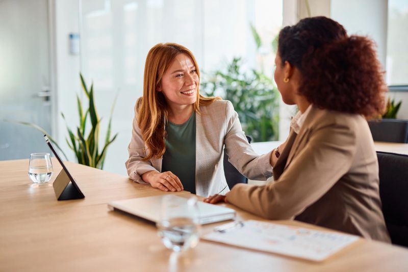 Two businesswomen sitting at a modern office table, engaging in a heartfelt conversation while providing support and comfort after a challenging workday, fostering teamwork and connection