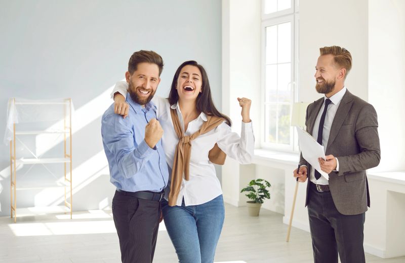 Happy married couple is joyfully and contentedly clenching their fists during meeting with real estate agent. Man and woman who just bought their dream home are laughing standing next to male real estate agent.