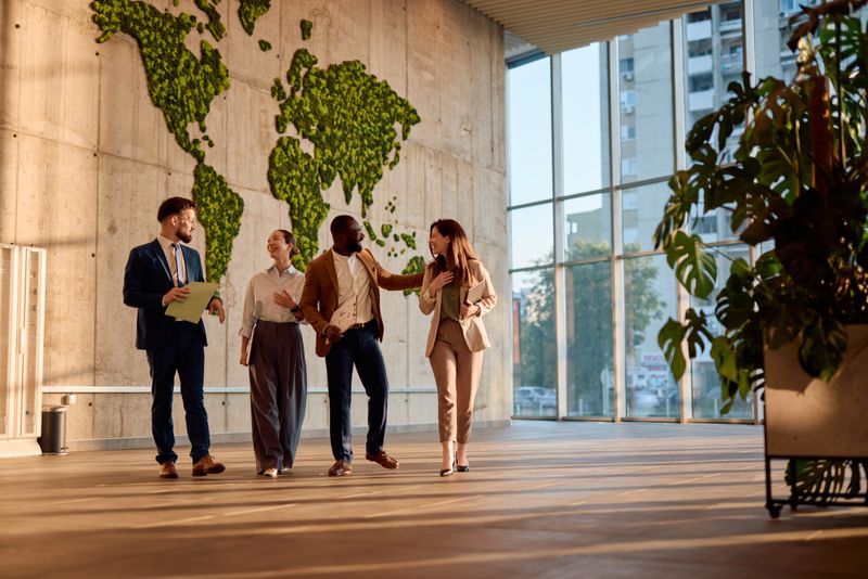 Four businesspeople walking and engaging in conversation within a modern office featuring a green world map, collaborating on global strategies and discussing sustainability initiatives