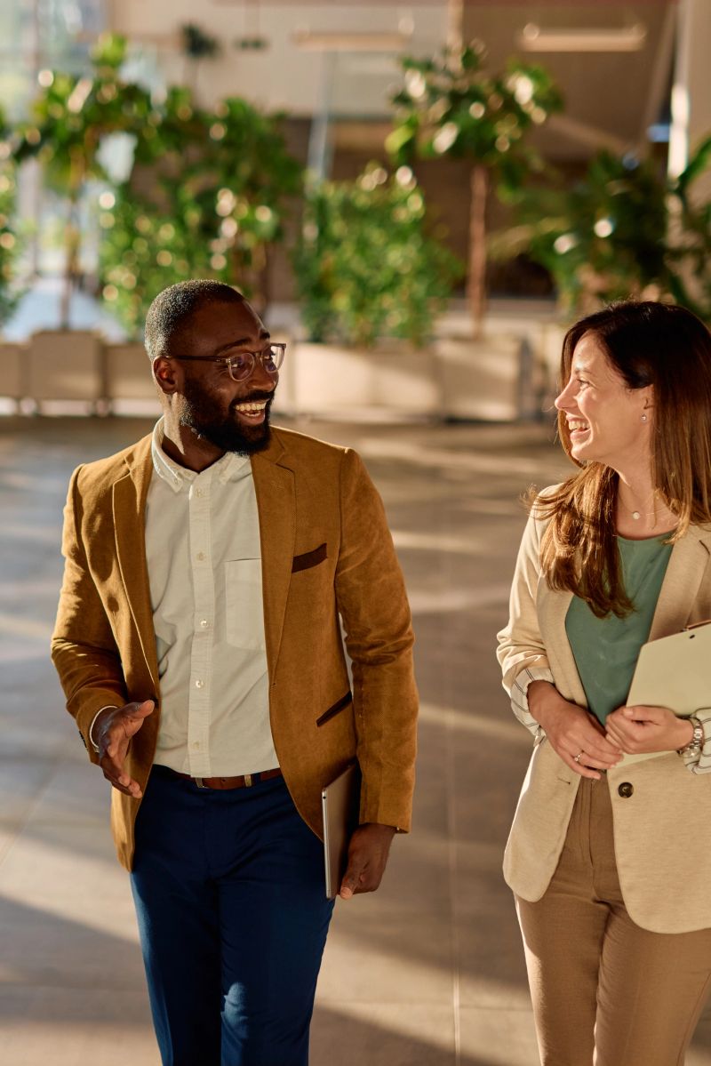 Cheerful businesspeople walking and engaging in conversation within a modern office building lobby, holding a tablet and clipboard while sharing smiles and ideas