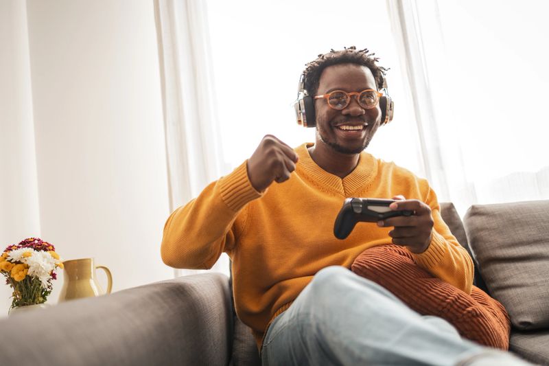 A cheerful man wearing headphones is seated on a cozy sofa, deeply engaged in a video game. His joyful expression reflects satisfaction and relaxation, capturing a moment of leisure and entertainment.