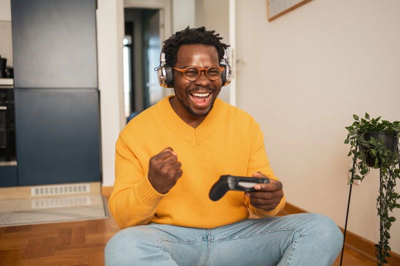 A joyful man celebrates a gaming success while wearing headphones and holding a controller at home. He is seated on the floor, radiating happiness and excitement in a relaxed environment.