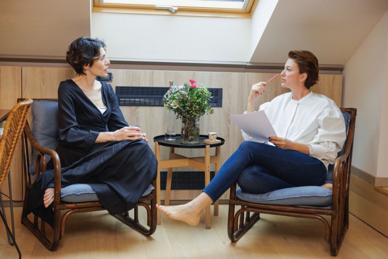 Two middle-aged women sitting in chairs by the roof window at home in the morning, reading and discussing menopause body care