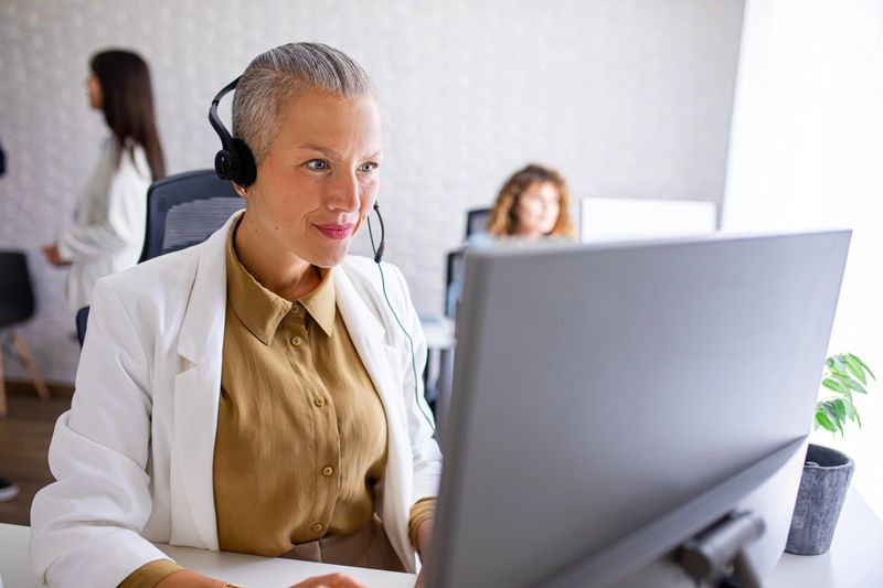 A friendly technical support manager is sitting at the office in call center and providing desk solutions to a customer while talking on hands free headset.