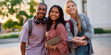Three diverse students smiling outdoors with books.