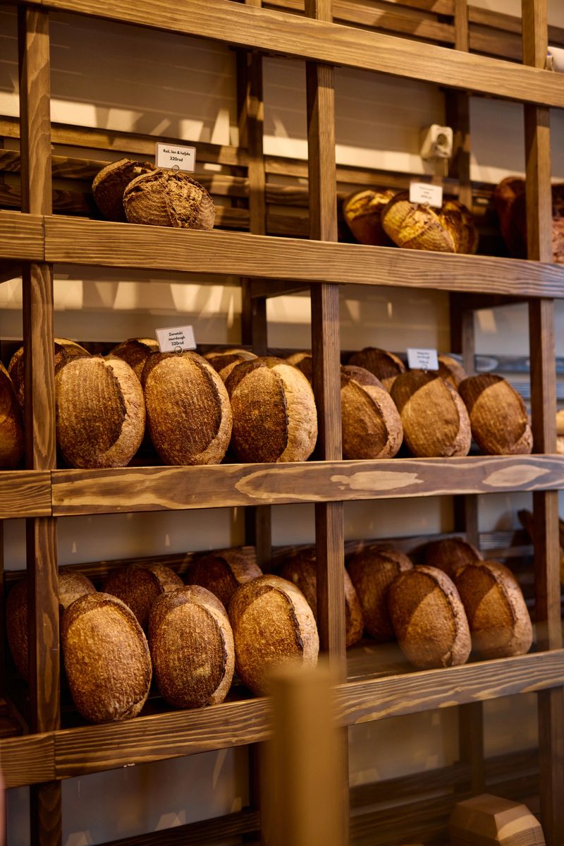 Freshly baked loaves of artisanal bread are neatly arranged on wooden shelves in a bakery display, showcasing a variety of textures and flavors for customers to choose from