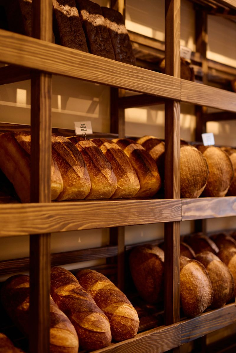 Warm lighting illuminates various types of freshly baked bread loaves arranged on wooden shelves, creating an inviting display in a bakery or artisanal bread shop