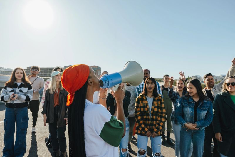 A diverse group of individuals demonstrating outdoors, with a woman leading through a megaphone. The event promotes community support for social causes and raising awareness for oppression.