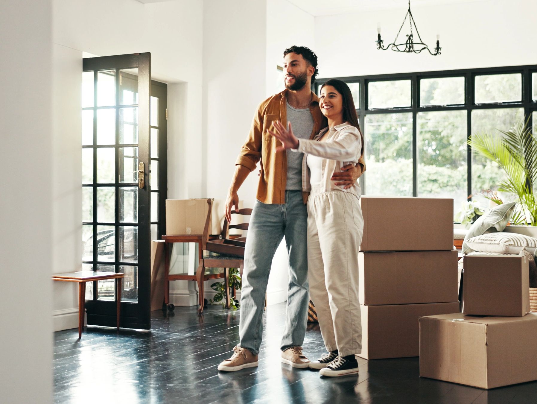 A smiling young couple stands inside their new, sunlit home surrounded by cardboard moving boxes. Th