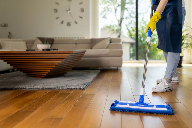 A person wearing yellow gloves uses a mop to wipe hardwood floors in a stylish living room. Sunlight streams through large windows, highlighting the organized space.