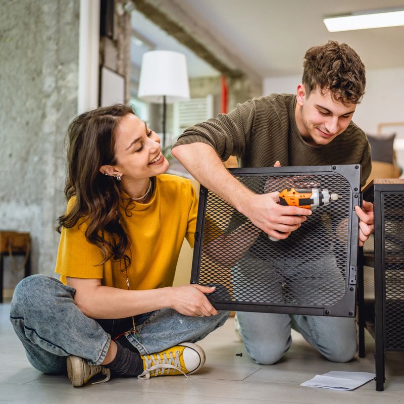 Adult couple have fun and use electric drill for assembling table at new apartment. Concept of family, relocation and manual work