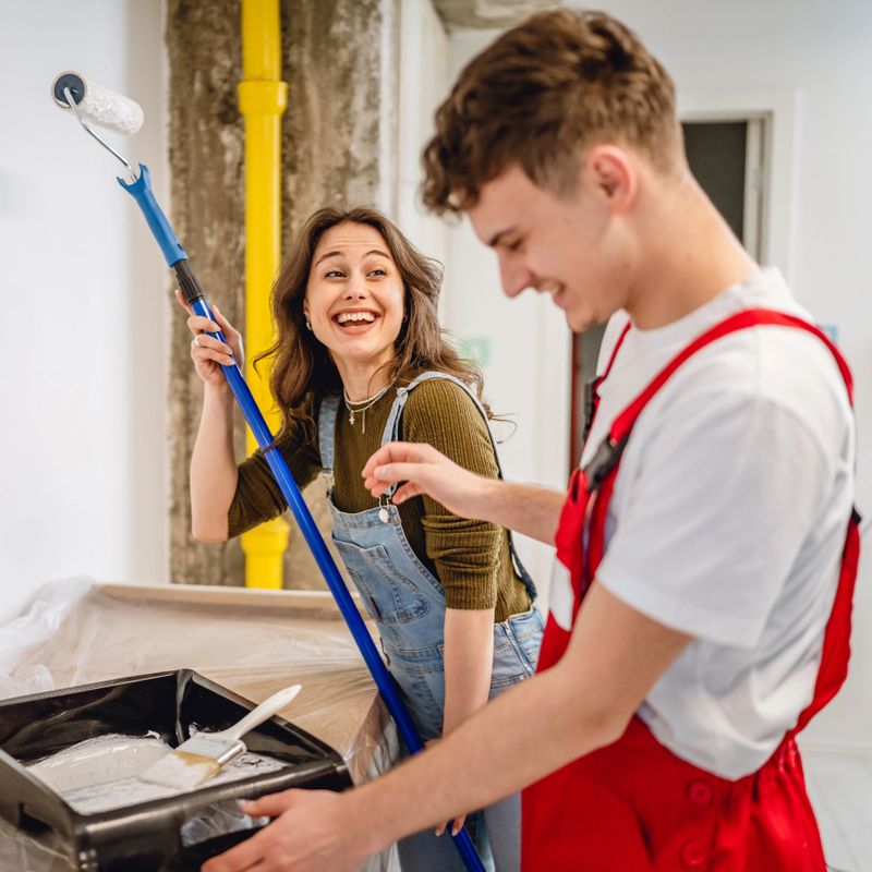 Happy young couple decorates home by painting wall in white color. Work together using paint rollers. Renovating house with love and enthusiasm