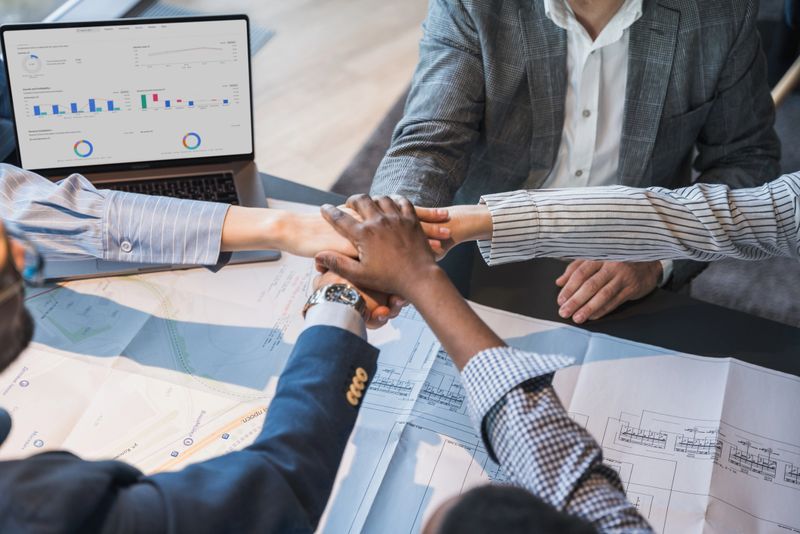 A group of professionals unite around a table, hands joined symbolically, surrounded by business documents, showcasing teamwork, collaboration, and shared commitment in a corporate environment.