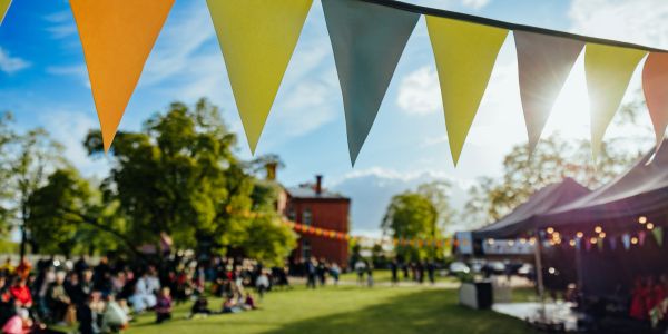 Colorful pennant flags hang above a lively outdoor festival with a crowd and stage.