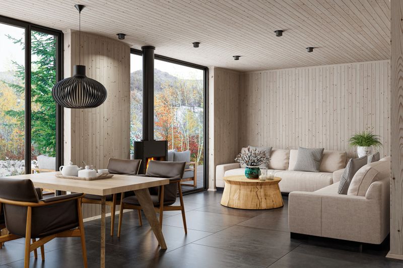 Warm and inviting Scandinavian-style living room captured from a new angle, showcasing two beige sofas arranged around a round solid-wood stump coffee table, decorated with minimalist candles for a cozy touch. The space features a freestanding modern black metal fireplace, a dining area with four chairs, and continuous pine wood paneling across the walls and ceiling. Matte black stone floor tiles add contrast and sophistication, enhancing the natural textures. Ideal for use in interior design promotions, eco-cabin marketing, Scandinavian lifestyle visuals, real estate, architectural presentations, and sustainable furniture or material branding.