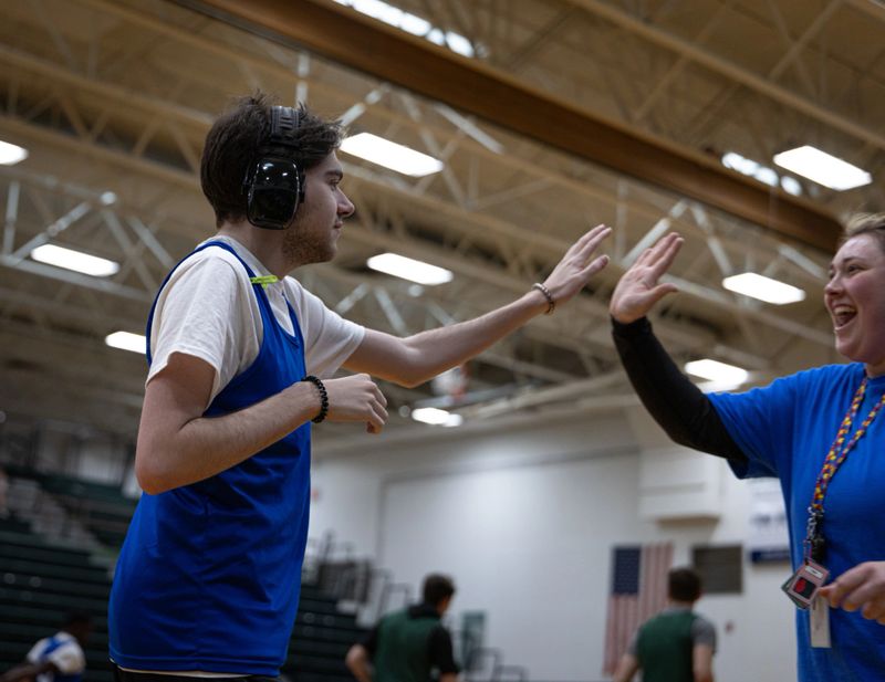 A 20 year old neurodivergent man with autism wearing noise canceling headphones high fives his female basketball coach before a game