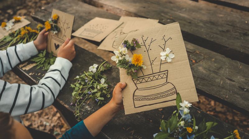Children creating handmade flower crafts using cardboard, drawing vases and attaching real spring blossoms. Creative outdoor activity on a rustic wooden table in the forest.