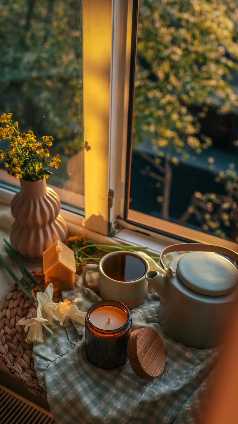 Cozy window nook with open book, ceramic vase of yellow flowers, cup of tea, teapot, and spring blossoms at sunset. Warm golden light creates a peaceful, reflective mood.