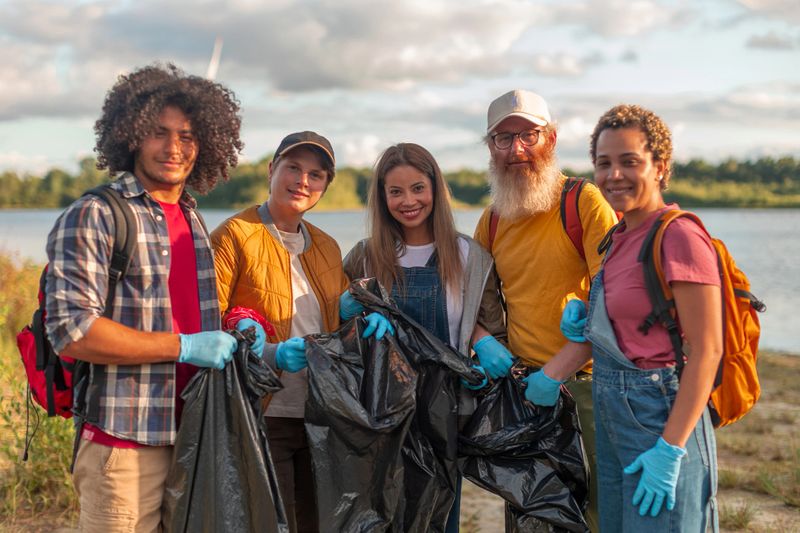 Group of volunteers holding trash bags, cleaning up garbage near lake, promoting environmental conservation and teamwork