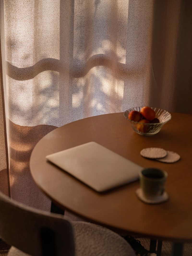 Intimate view of a wooden round table with closed laptop and ceramic mug on a woven coaster. A glass bowl of tangerines adds vibrant color against the warm oak surface. Translucent curtains filter sunlight, casting ethereal tree shadow patterns across the peaceful morning scene.