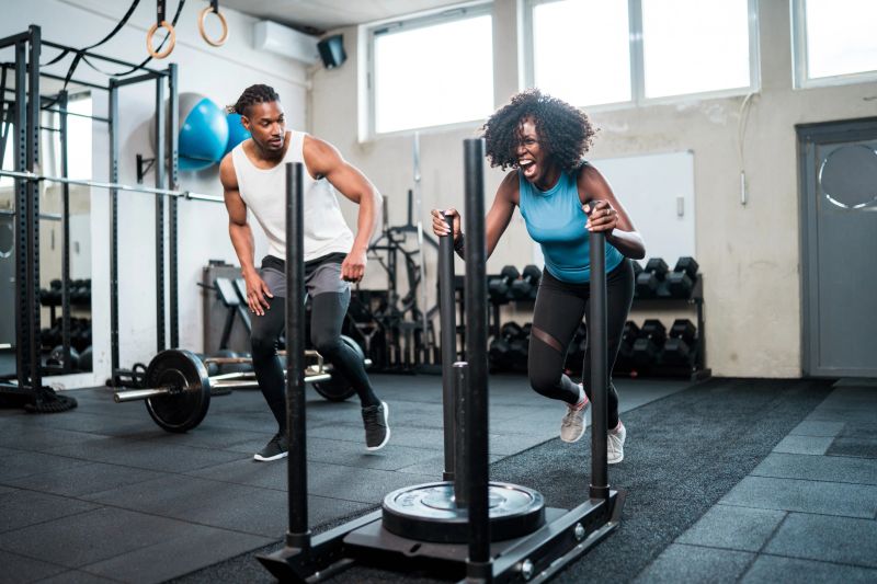 A mid adult Black woman pushes a weighted sled in a modern gym, guided by a focused male trainer in athletic attire, embodying strength and determination.