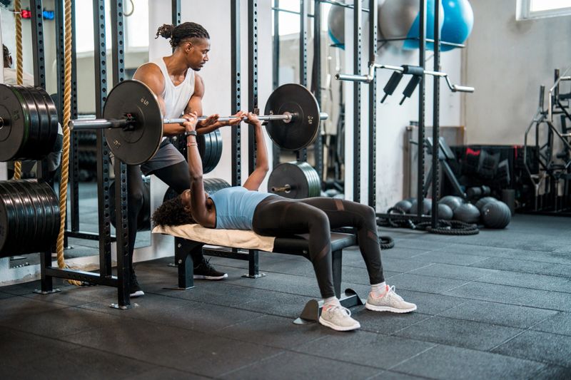 A mid adult Black female bench presses weights under the guidance of a personal trainer in a bright, modern gym. The scene is energetic, with fitness equipment and weights in the background, emphasizing strength and determination.