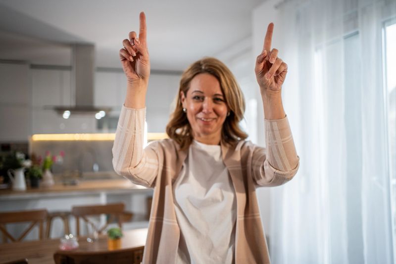 Mid adult woman smiles and points upwards with both her index fingers while standing in her modern kitchen, suggesting or highlighting something above
